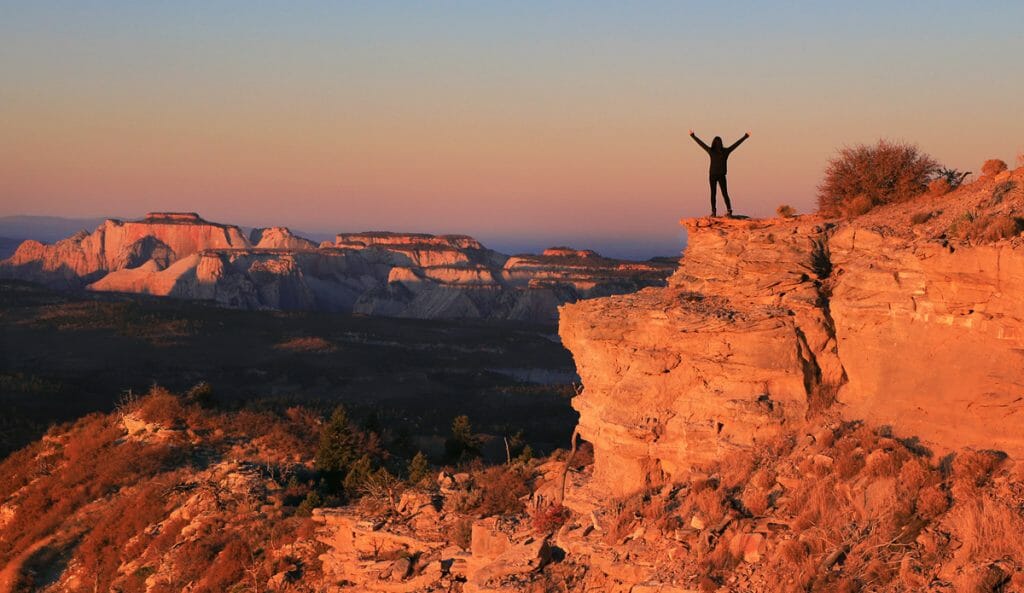 A person stands triumphantly on a rock ledge overlooking a vast landscape at sunset.